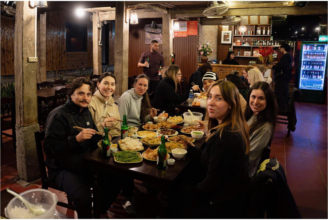 A group of diverse international travelers sitting at a long wooden table in a traditional rustic restaurant, enjoying an authentic Vietnamese meal with chopsticks and local beer. The warm, communal atmosphere highlights the culinary side of cultural exchange and ethnic diversity