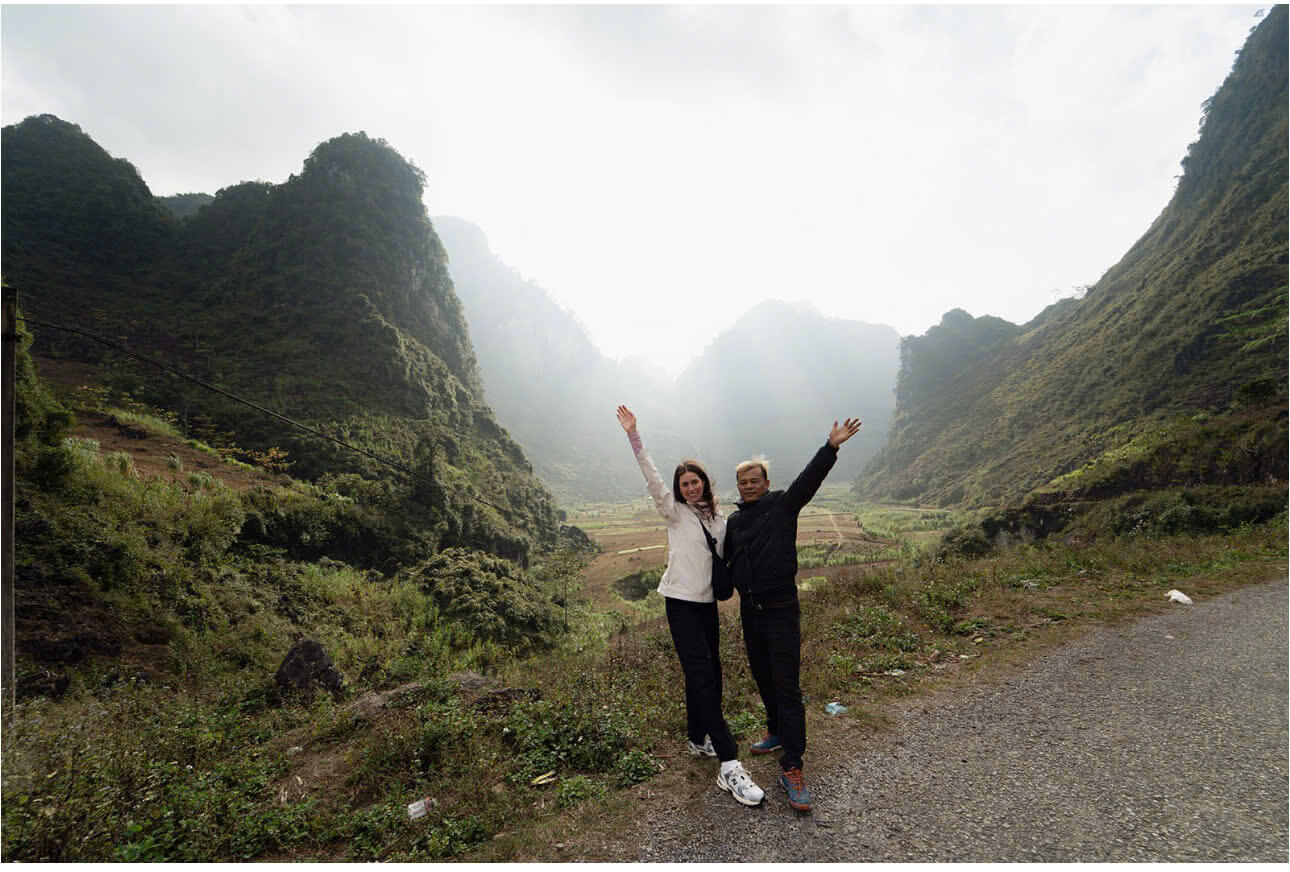 A man and woman waving their hands on a roadside in a dramatic, misty mountain valley, showcasing the atmospheric winter weather conditions on the Ha Giang Loop.