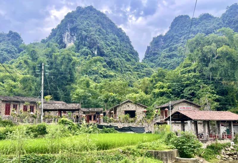 A scenic view of the Khuoi Ky stone village, featuring traditional houses made of local stone with tiled roofs, nestled at the foot of lush, green limestone mountains.