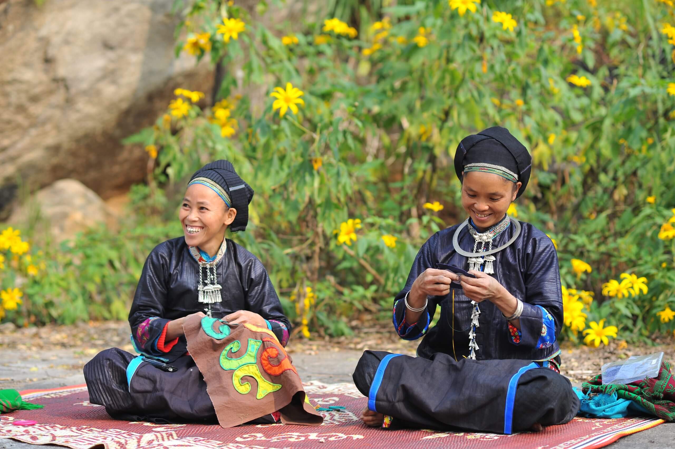 Two ethnic Nung women in traditional indigo clothing practicing embroidery outdoors on the Ha Giang Loop, Vietnam.