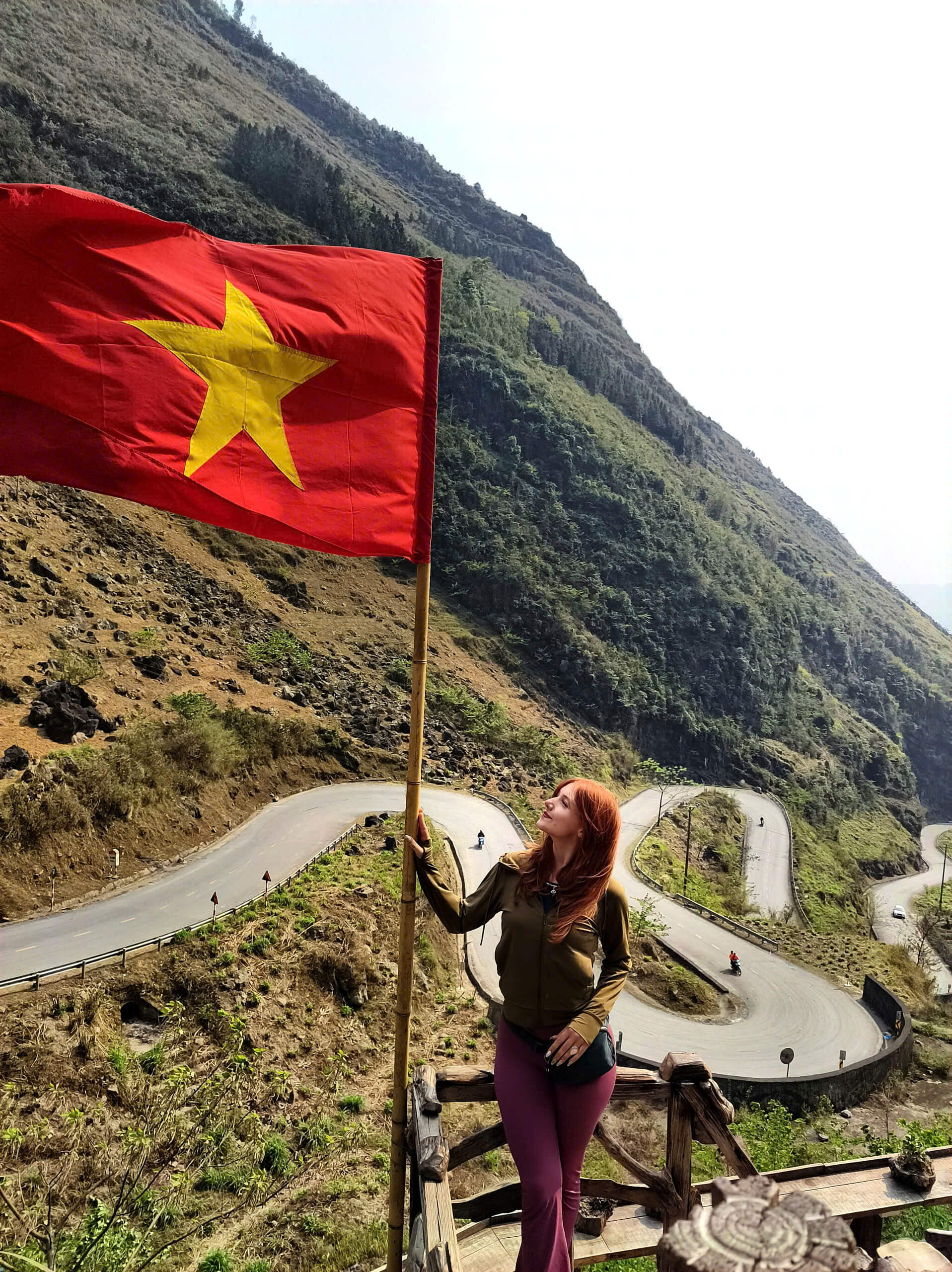 A solo female traveler standing by the Vietnamese flag at Tham Ma Pass, enjoying the breathtaking scenery of the untamed Ha Giang Loop route.