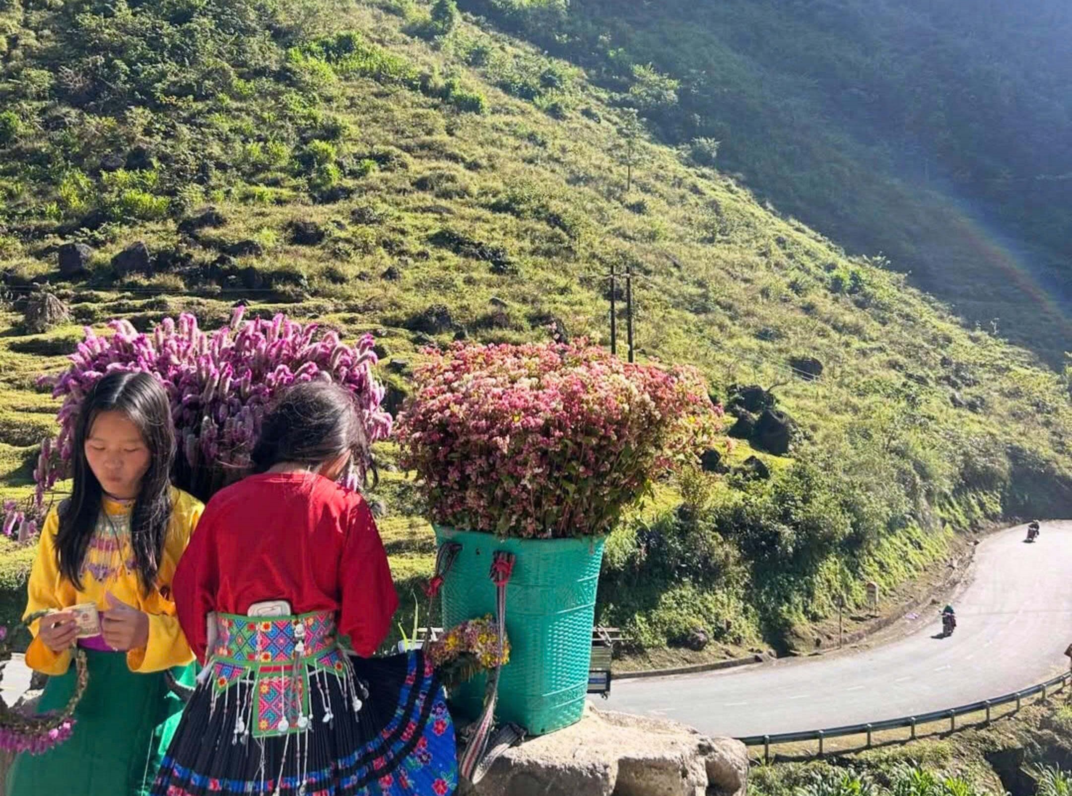 Ethnic children standing with flower baskets near a winding mountain road, showing the reality of local life on the Ha Giang Loop.