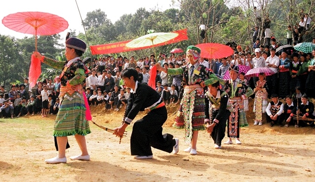 A wide shot of the Ha Giang festival grounds featuring a traditional tall ritual pole decorated with flags and a large crowd of attendees.