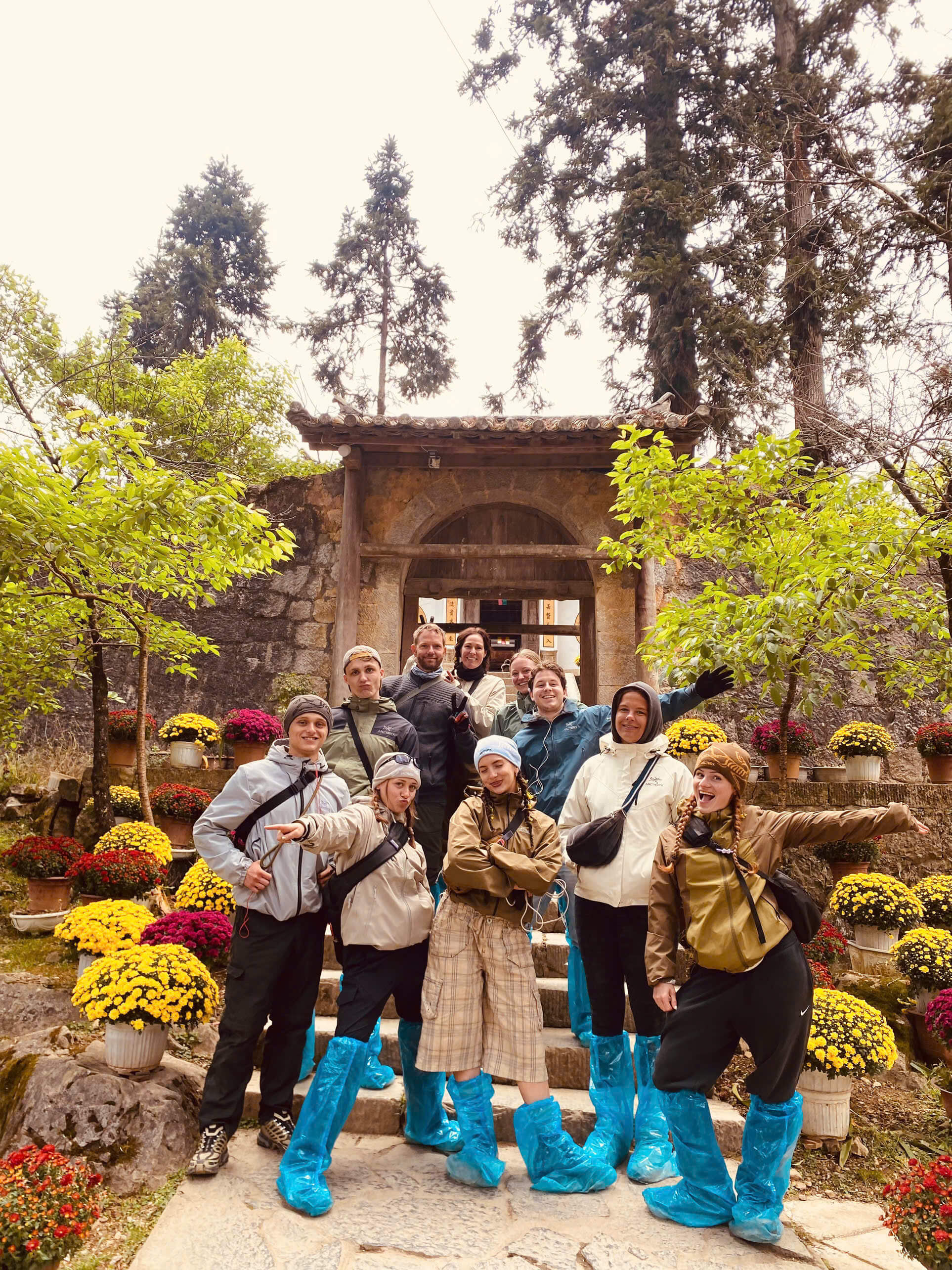 Travelers wearing protective rain shoe covers visiting a historical Hmong mansion decorated with blooming flowers during their spring trip in Ha Giang.
