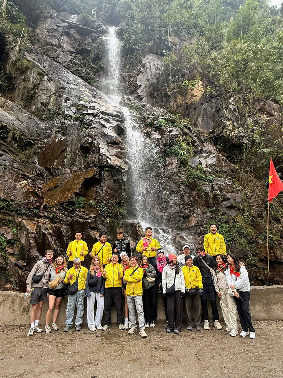 Tourists and Ha Giang Motorventures Easy Rider guides in yellow jackets taking a group photo in front of a beautiful waterfall on the Ha Giang Loop