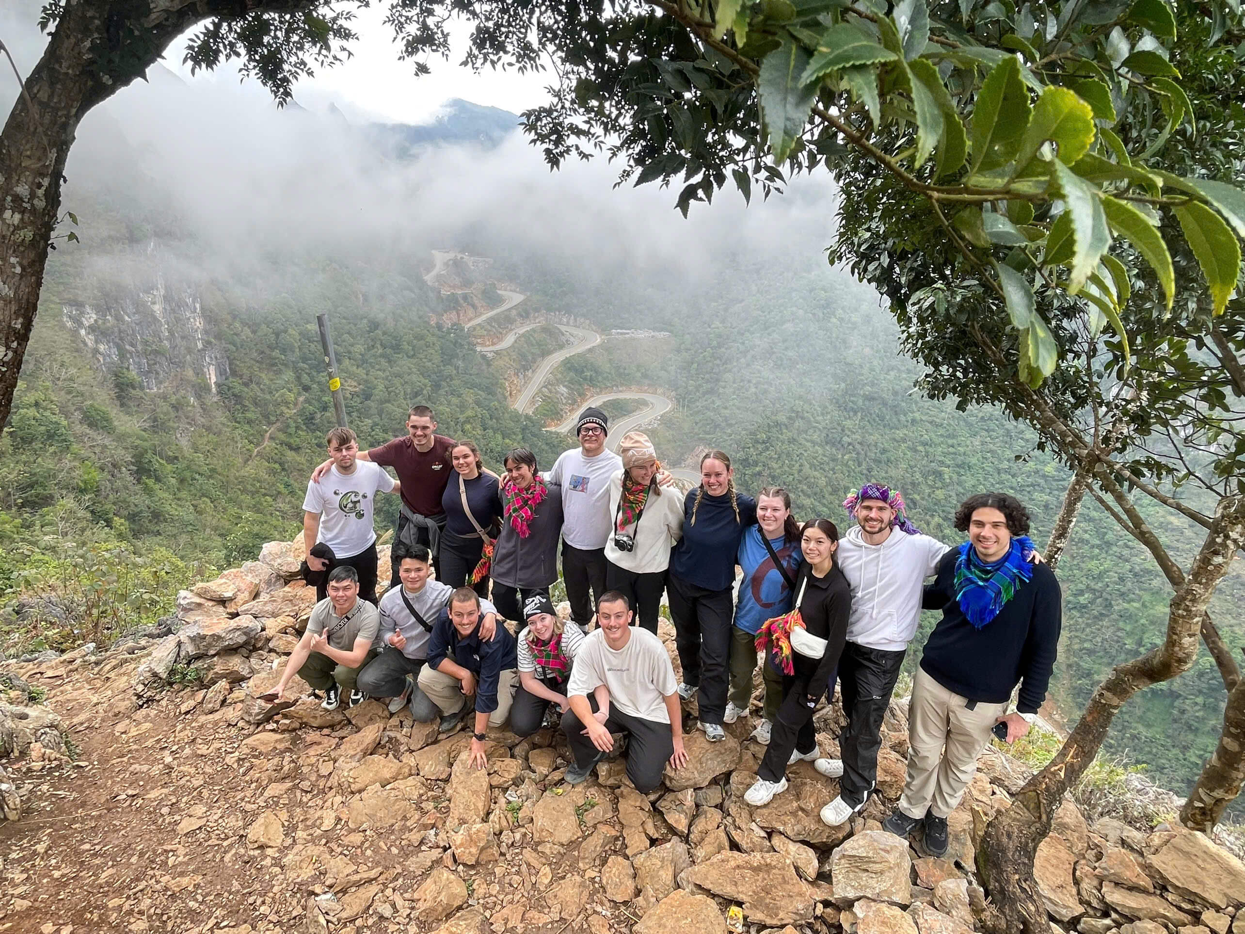 A happy group of backpackers and local guides from Ha Giang Motorventures posing on a rocky cliff overlooking a majestic, winding mountain pass.