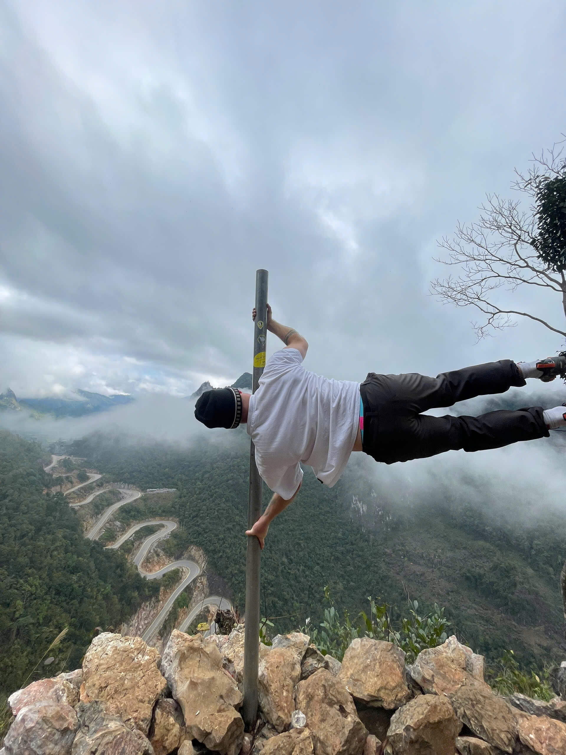 An adventurous traveler doing a human flag pole trick on the edge of a cliff with spectacular winding roads in the background during a motorbike tour.